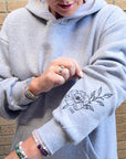 Person wearing a gray hoodie with a floral design on the sleeve, standing against a brick wall.