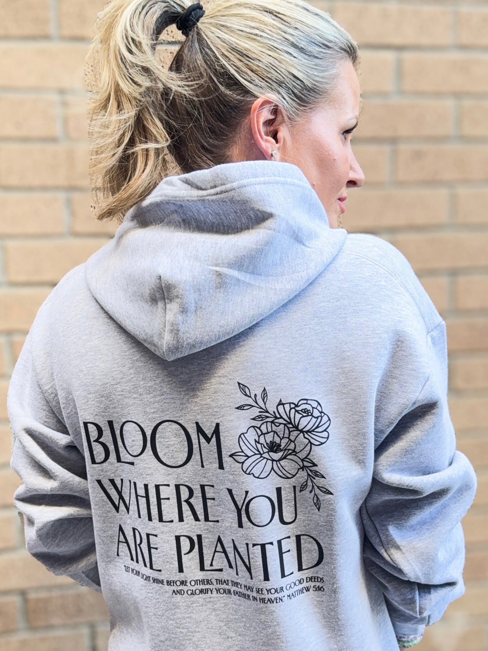 Person wearing a gray hoodie with 'Bloom Where You Are Planted' text and floral design on the back, standing against a brick wall.