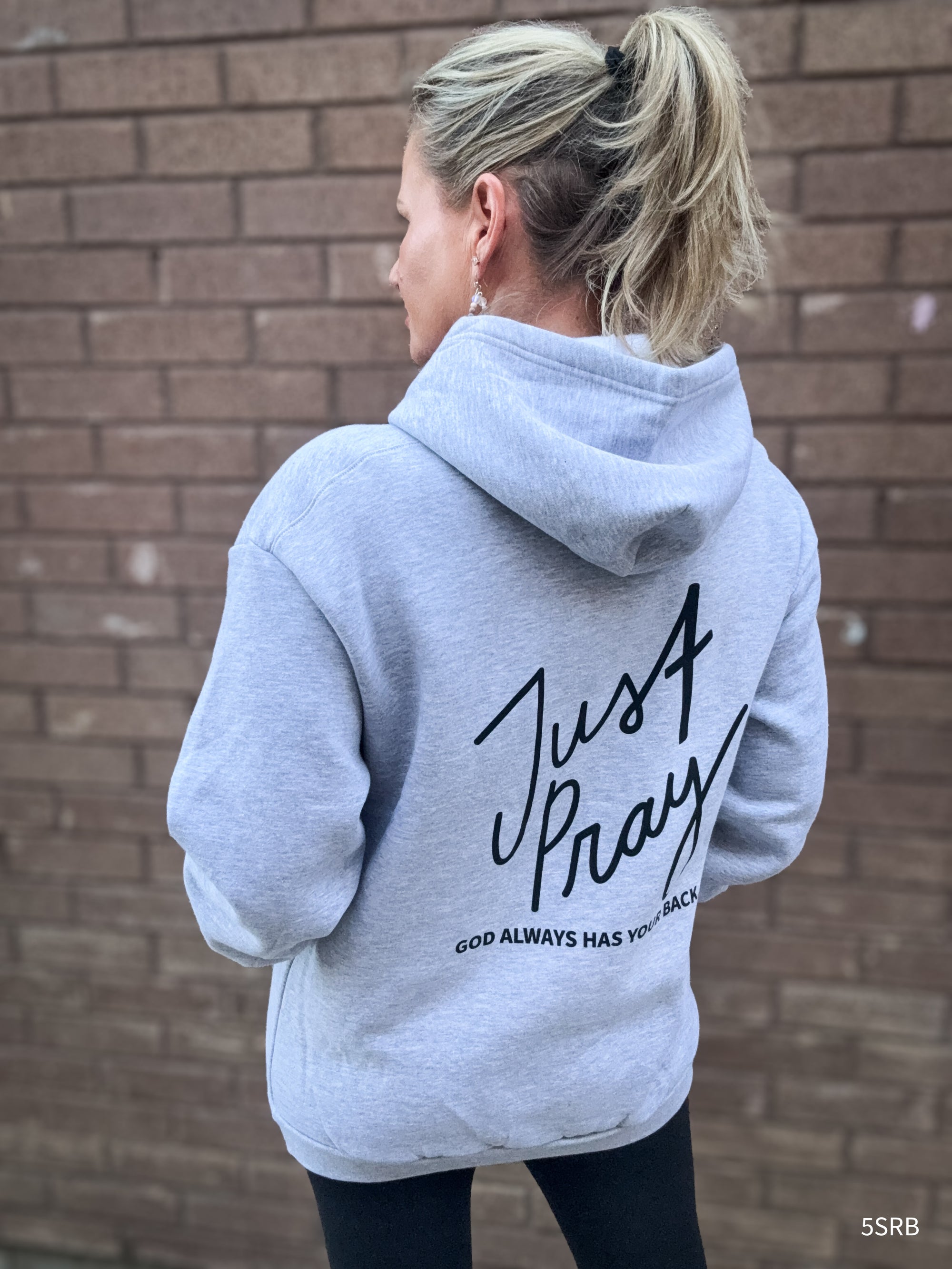 Person wearing a gray hoodie with 'Just Pray' text on the back against a brick wall.