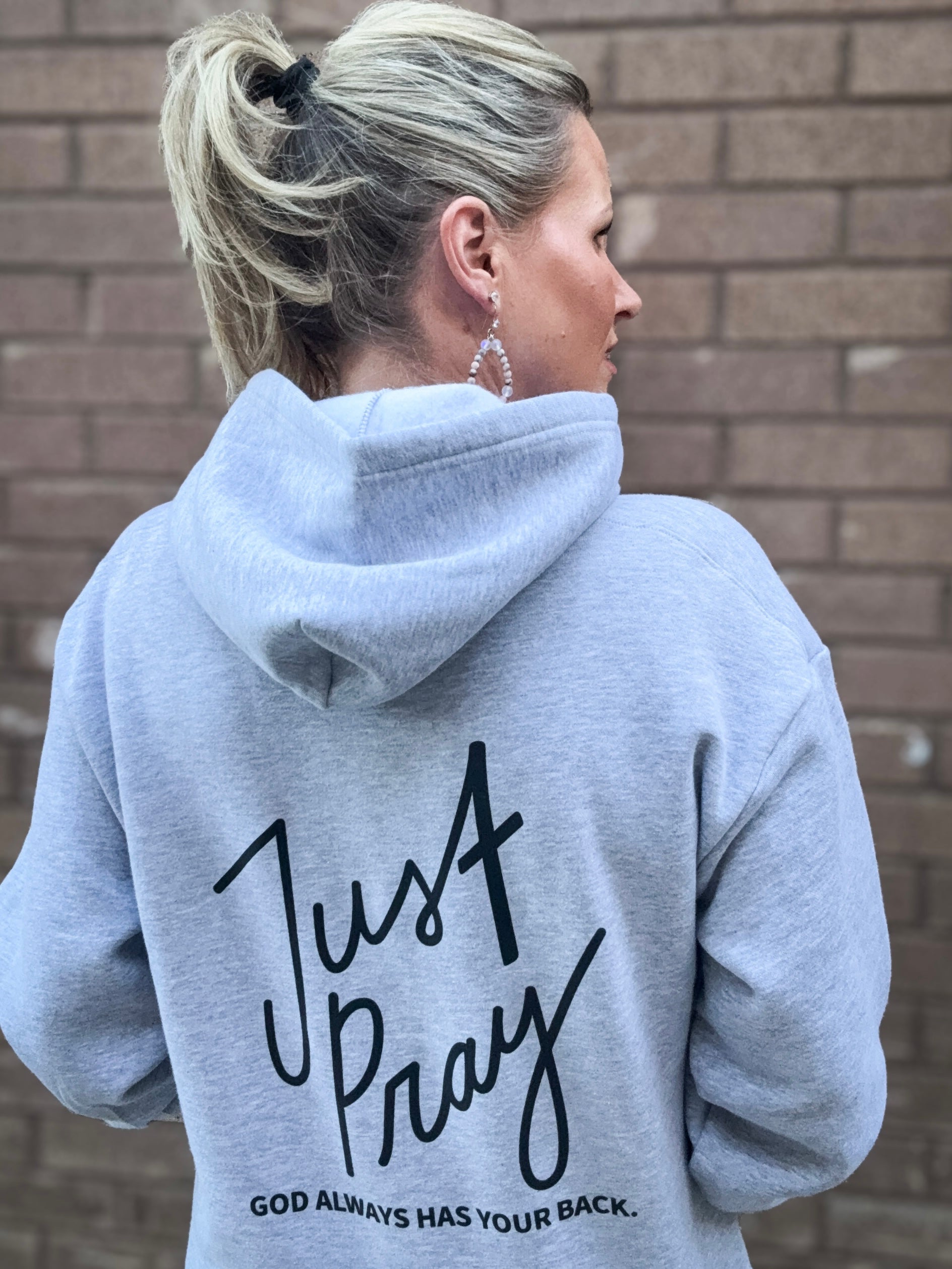 Person wearing a gray hoodie with 'Just Pray' text on the back against a brick wall.