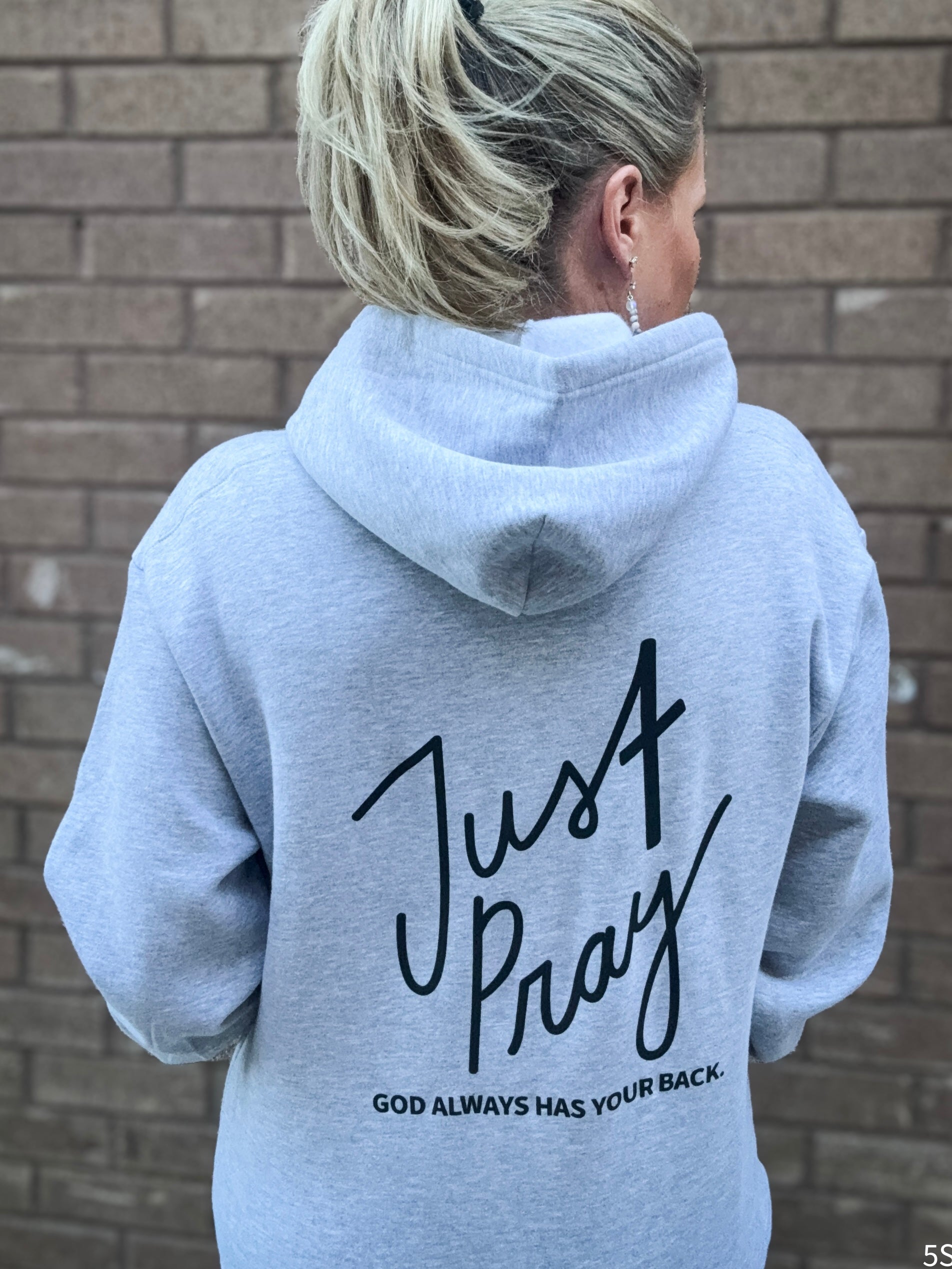 Person wearing a gray hoodie with 'Just Pray' text on the back against a brick wall.