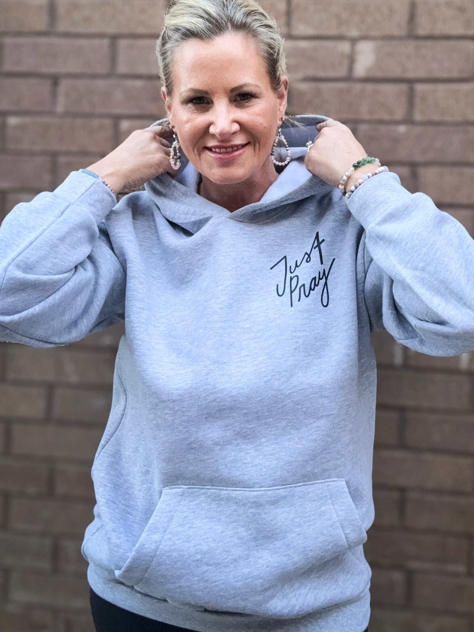 Person wearing a gray hoodie with 'Just Pray' text, standing against a brick wall.