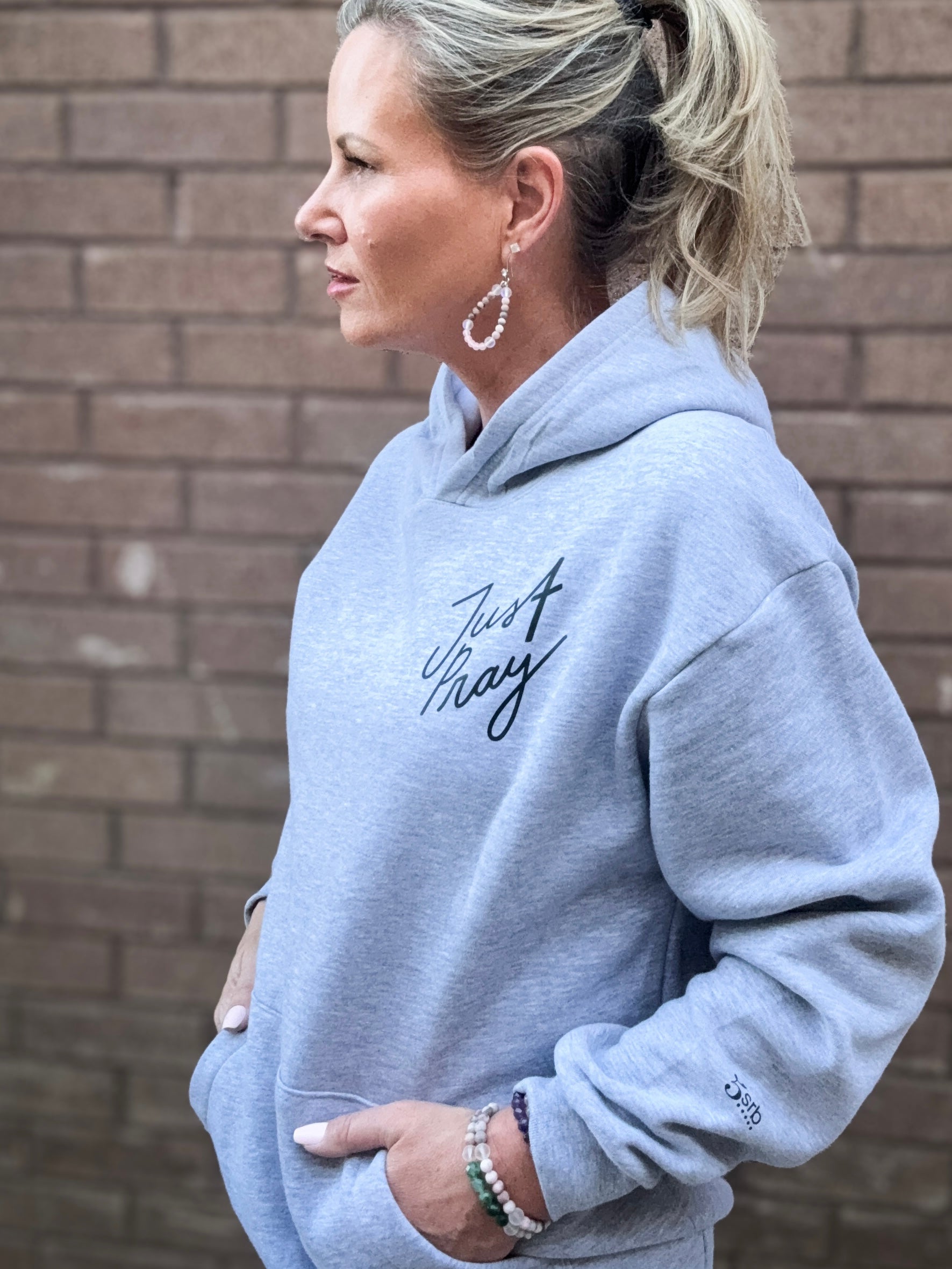 Woman wearing a light gray hoodie with text, standing against a brick wall.