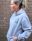 Woman wearing a light gray hoodie with text, standing against a brick wall.