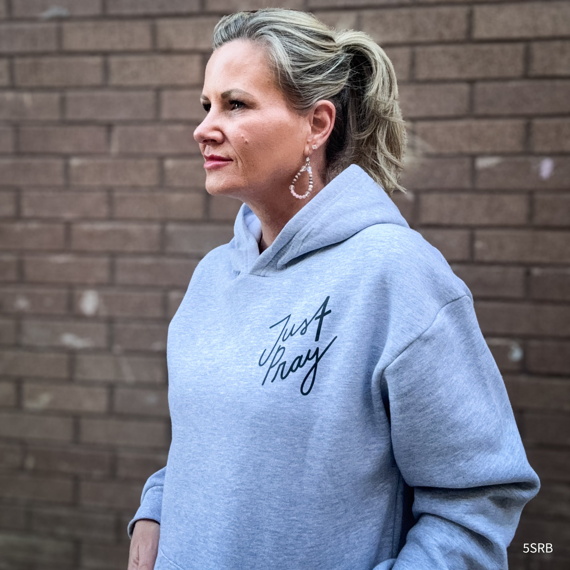 Woman wearing a gray hoodie with 'Just Stay' text against a brick wall.