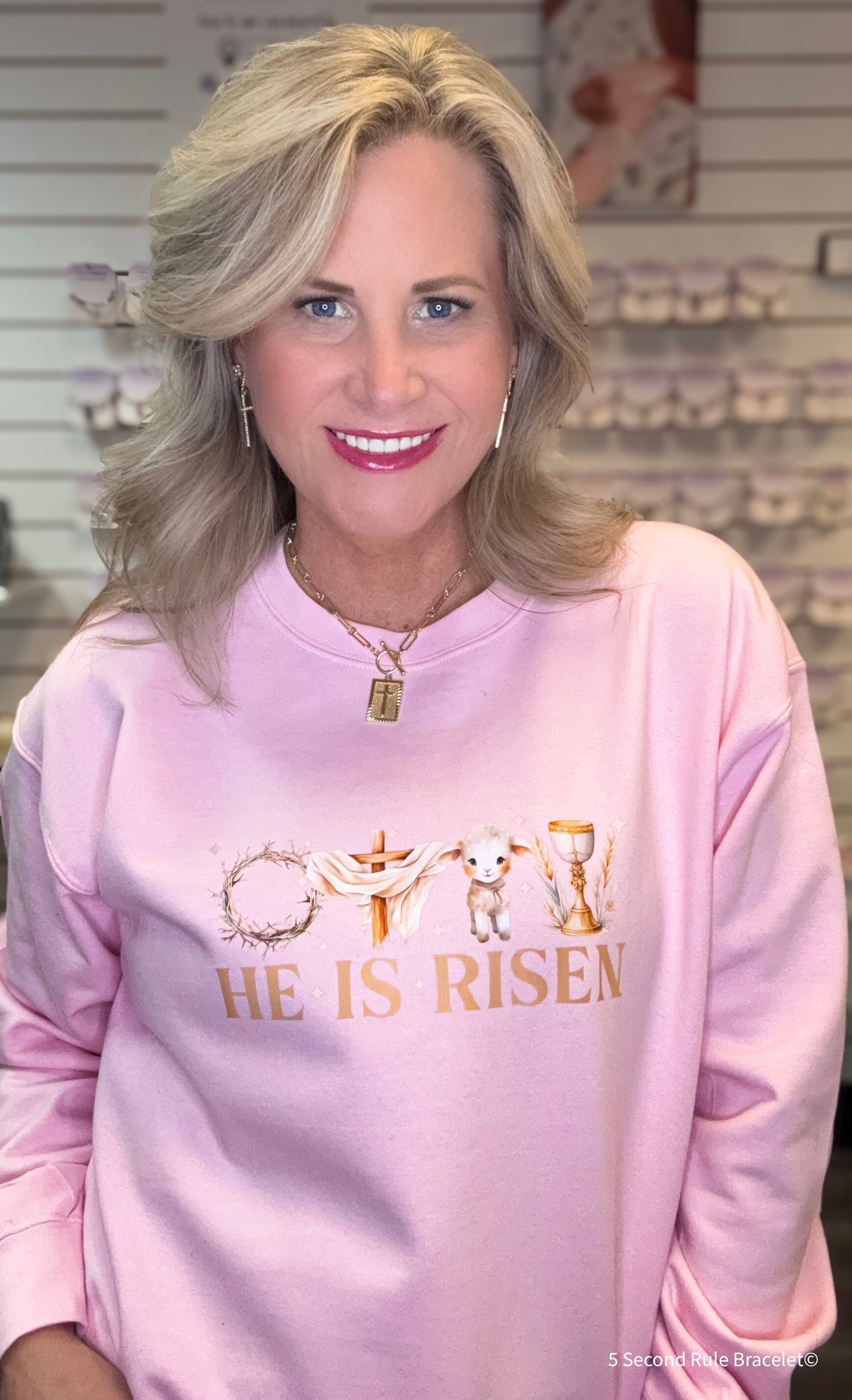 Woman wearing a pink sweatshirt with 'He is Risen' text and cross design.