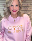Woman wearing a pink sweatshirt with 'He is Risen' text and cross design.
