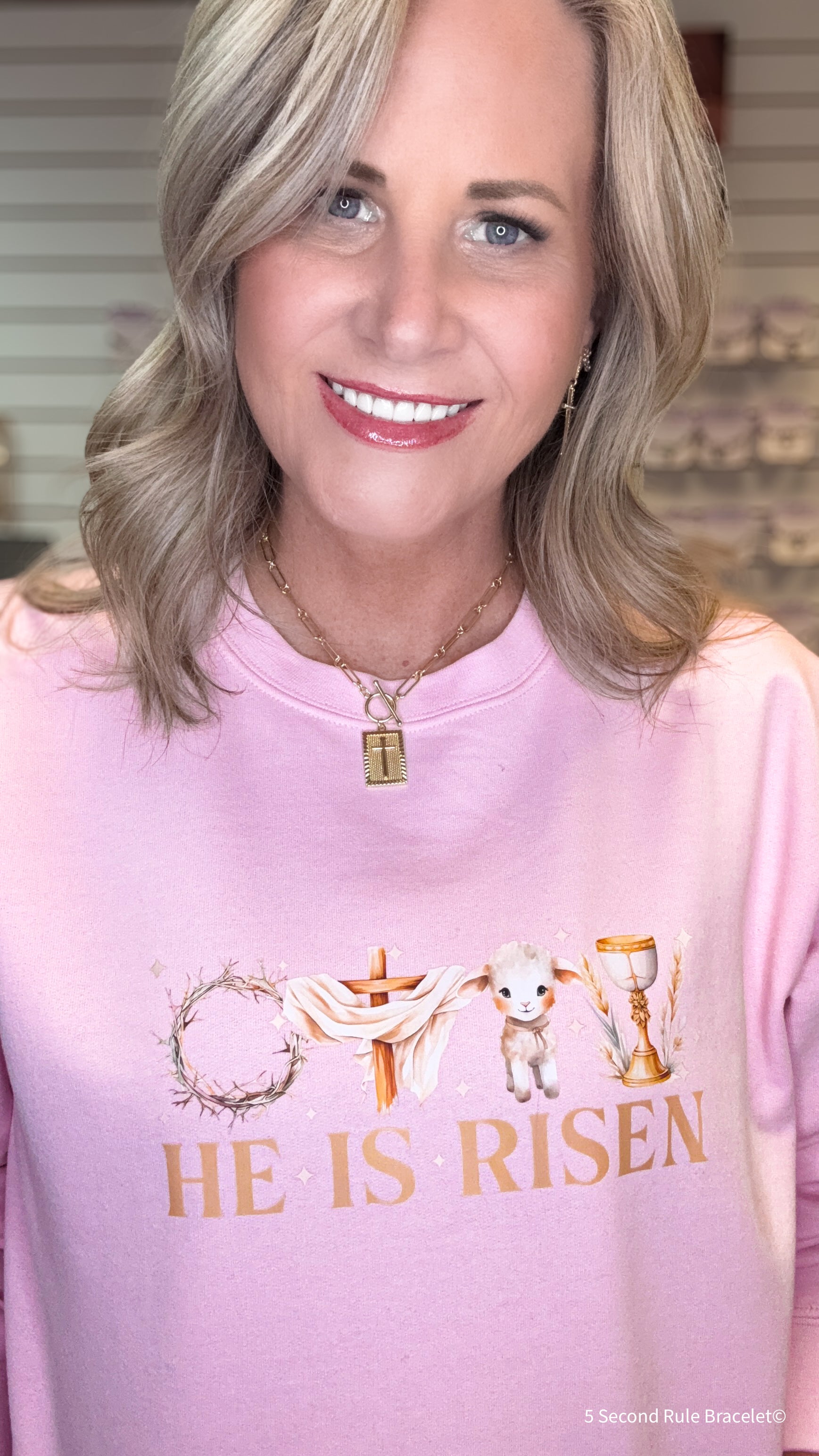 Woman wearing a pink sweatshirt with 'He is Risen' text and religious symbols.