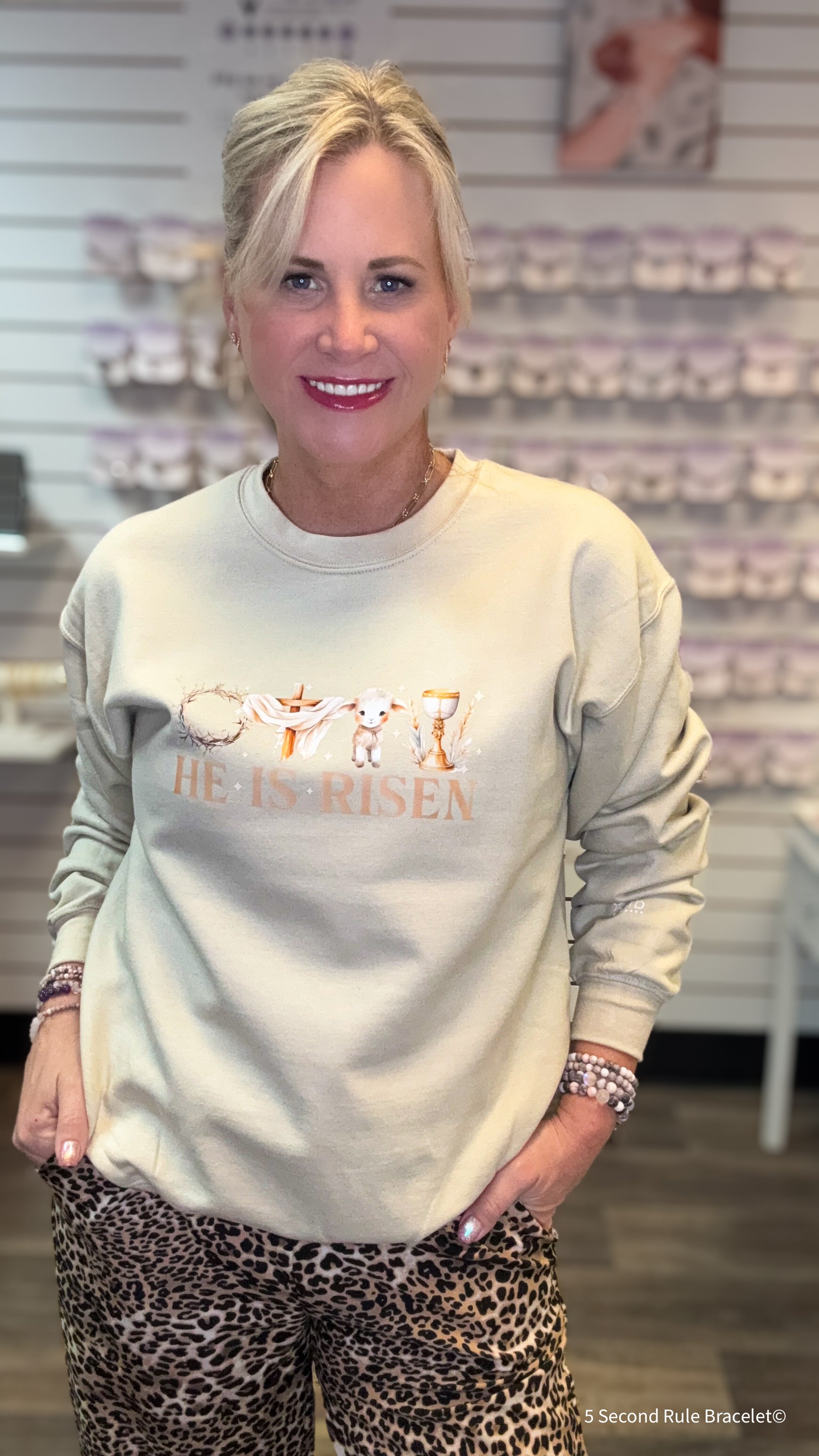Woman wearing a light-colored sweatshirt with 'He is Risen' text and leopard print pants in a store setting.