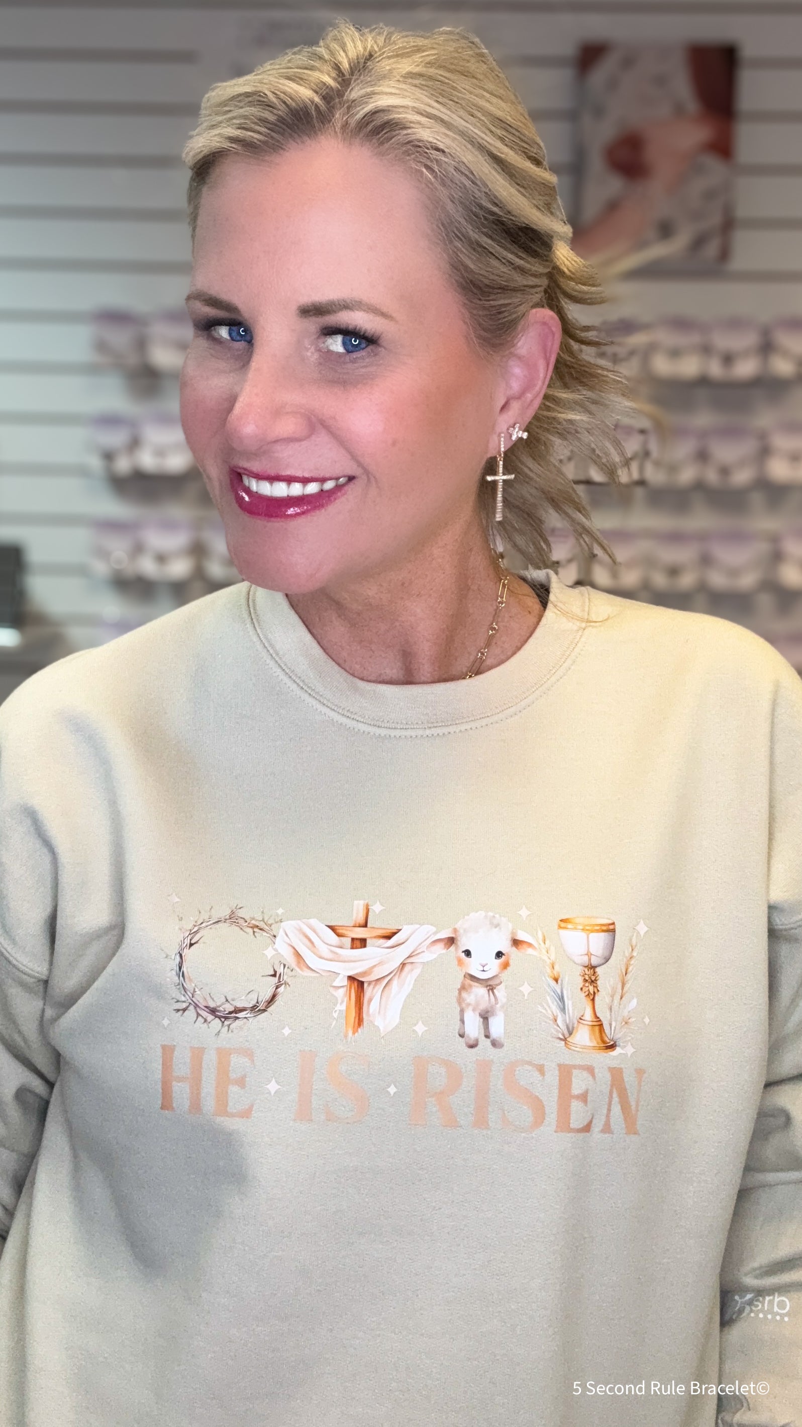 Woman wearing a light gray sweatshirt with 'He is Risen' text and cross design.