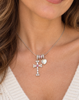 A close-up of a woman wearing a delicate silver cross necklace with pink gemstone accents and a small silver heart charm, paired with a white knit top.