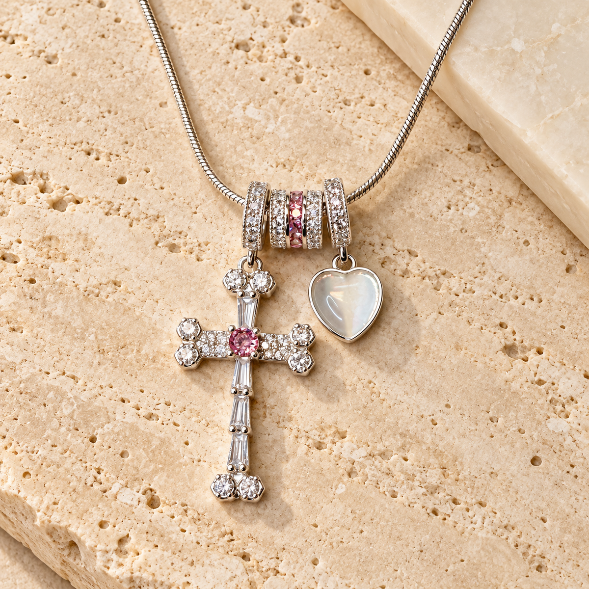 A silver-toned beaded necklace with a cross charm, a heart charm, and pink crystal accents, displayed on a textured beige stone surface.