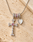 A silver-toned beaded necklace with a cross charm, a heart charm, and pink crystal accents, displayed on a textured beige stone surface.