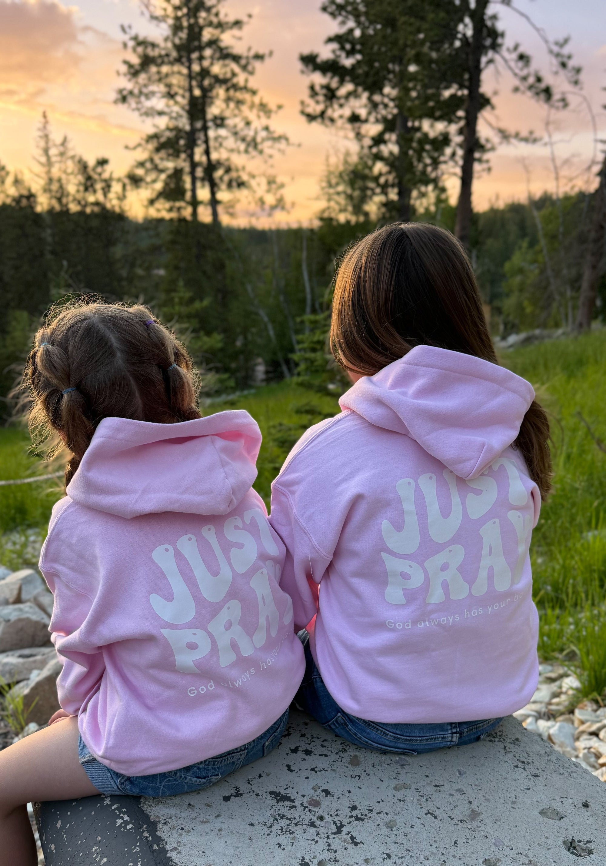 Two children wearing pink hoodies with &#39;JUST PRAY&#39; printed on the back, sitting on a rock at sunset.