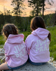 Two children wearing pink hoodies with 'JUST PRAY' printed on the back, sitting on a rock at sunset.