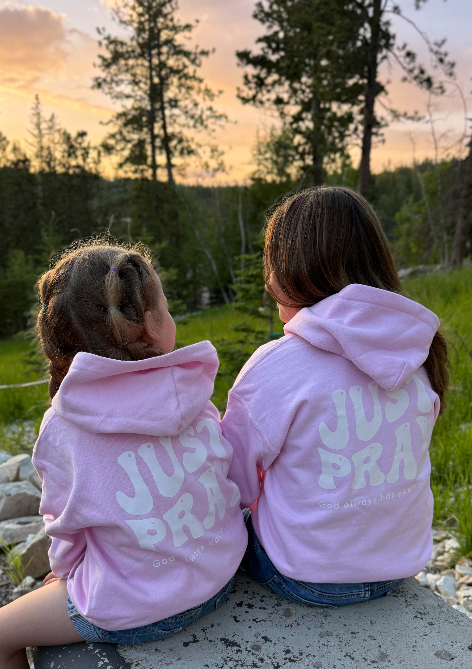 Two children wearing pink hoodies with 'Just Pray' text, sitting on a rock in a natural setting.