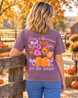 A woman viewed from behind wearing a heathered mauve-purple graphic t-shirt with a fall harvest design featuring pumpkins, flowers, and the text 'Jesus is the reason for the season'. She is standing outdoors on a rustic wooden fence in an autumn farm setting with pumpkins and fall foliage.