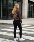 Woman in a brown sweatshirt and black pants crossing a street.