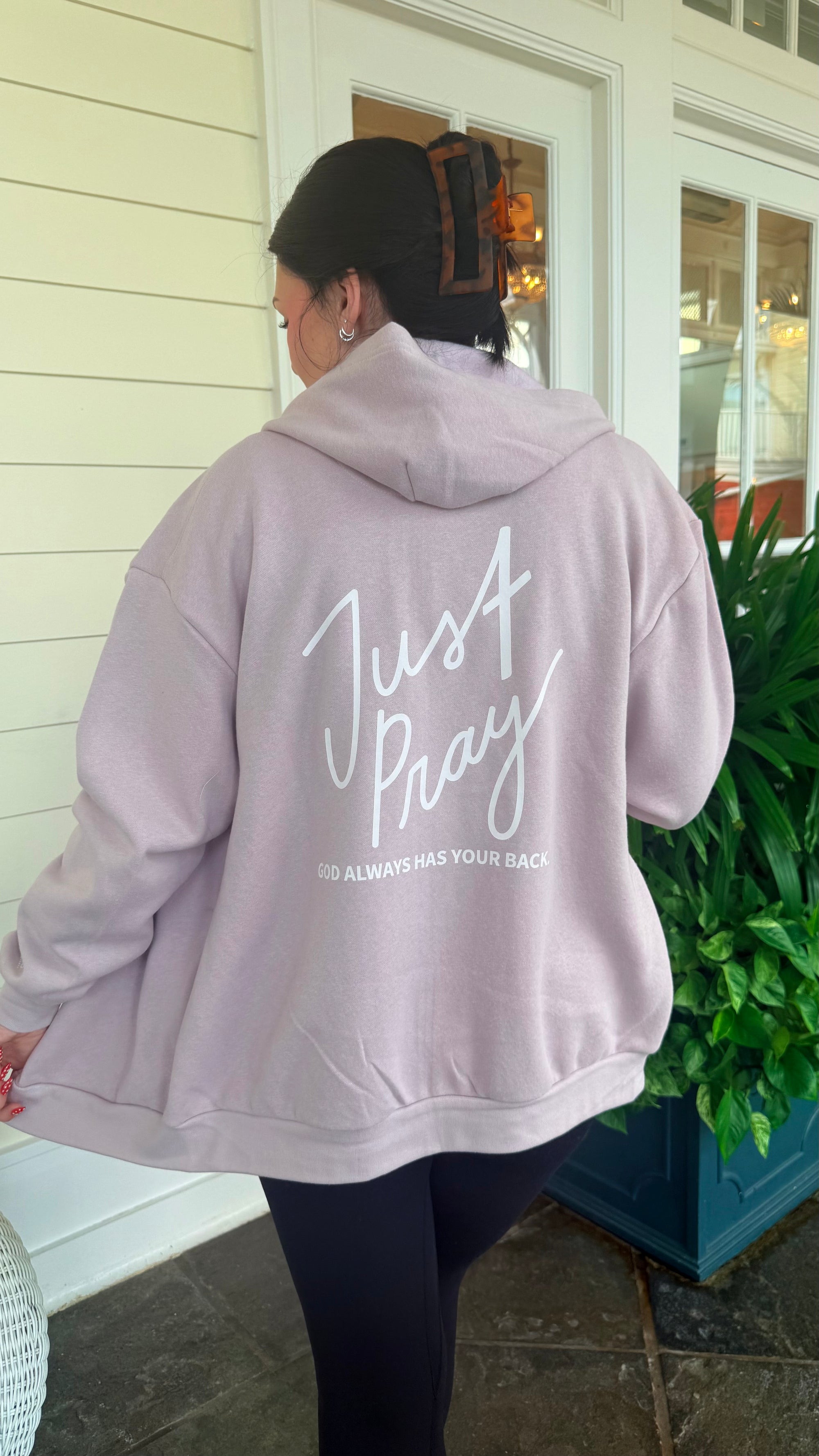 Person wearing a light pink hoodie with 'Just Pray' text on the back, standing outside.