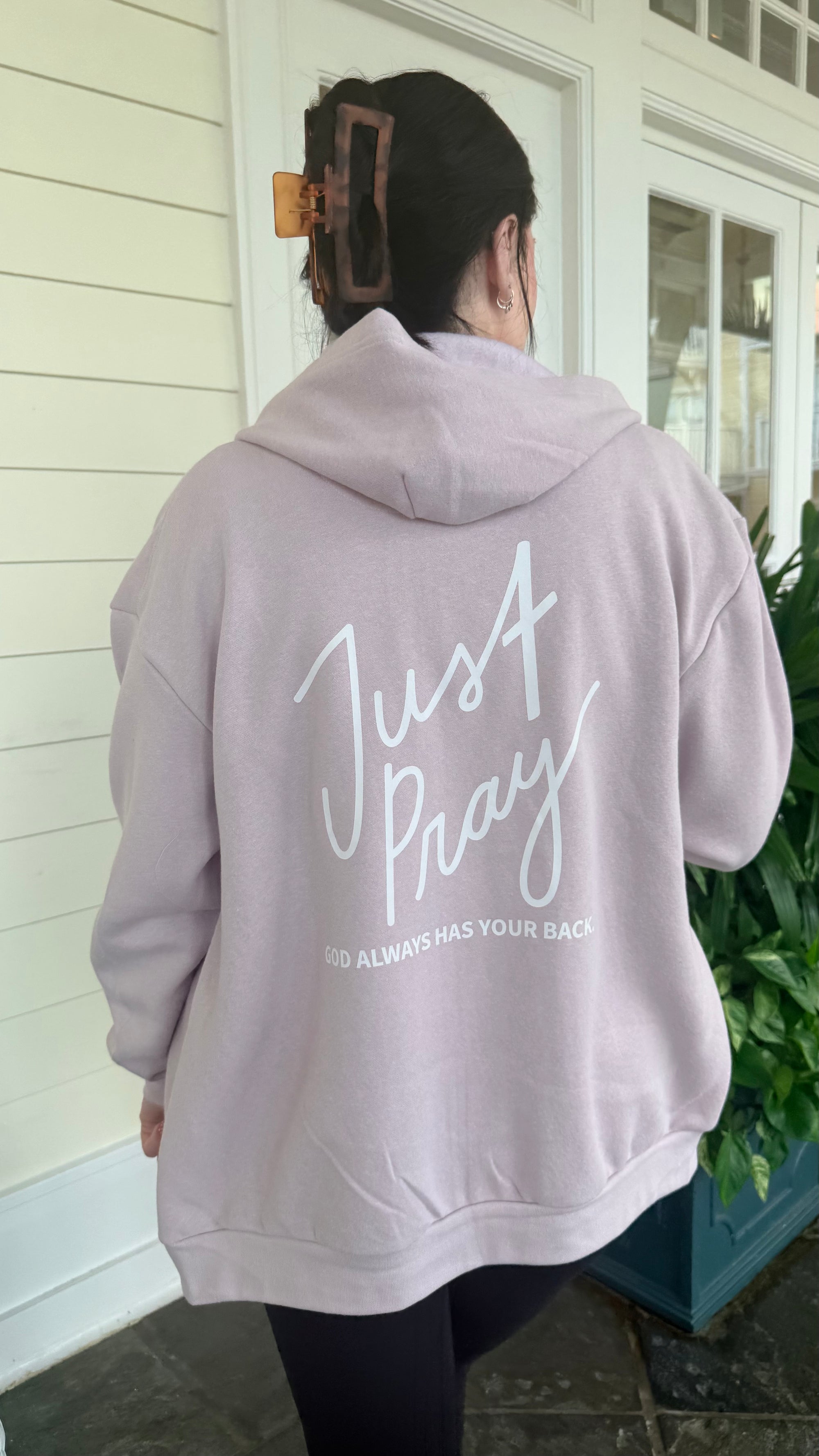 Person wearing a light pink hoodie with 'Just Pray' text on the back, standing outside.