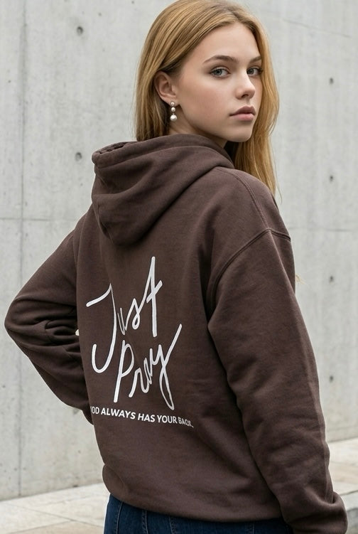 Person wearing a brown hoodie with 'Just Pray' text on the back, standing against a concrete wall.