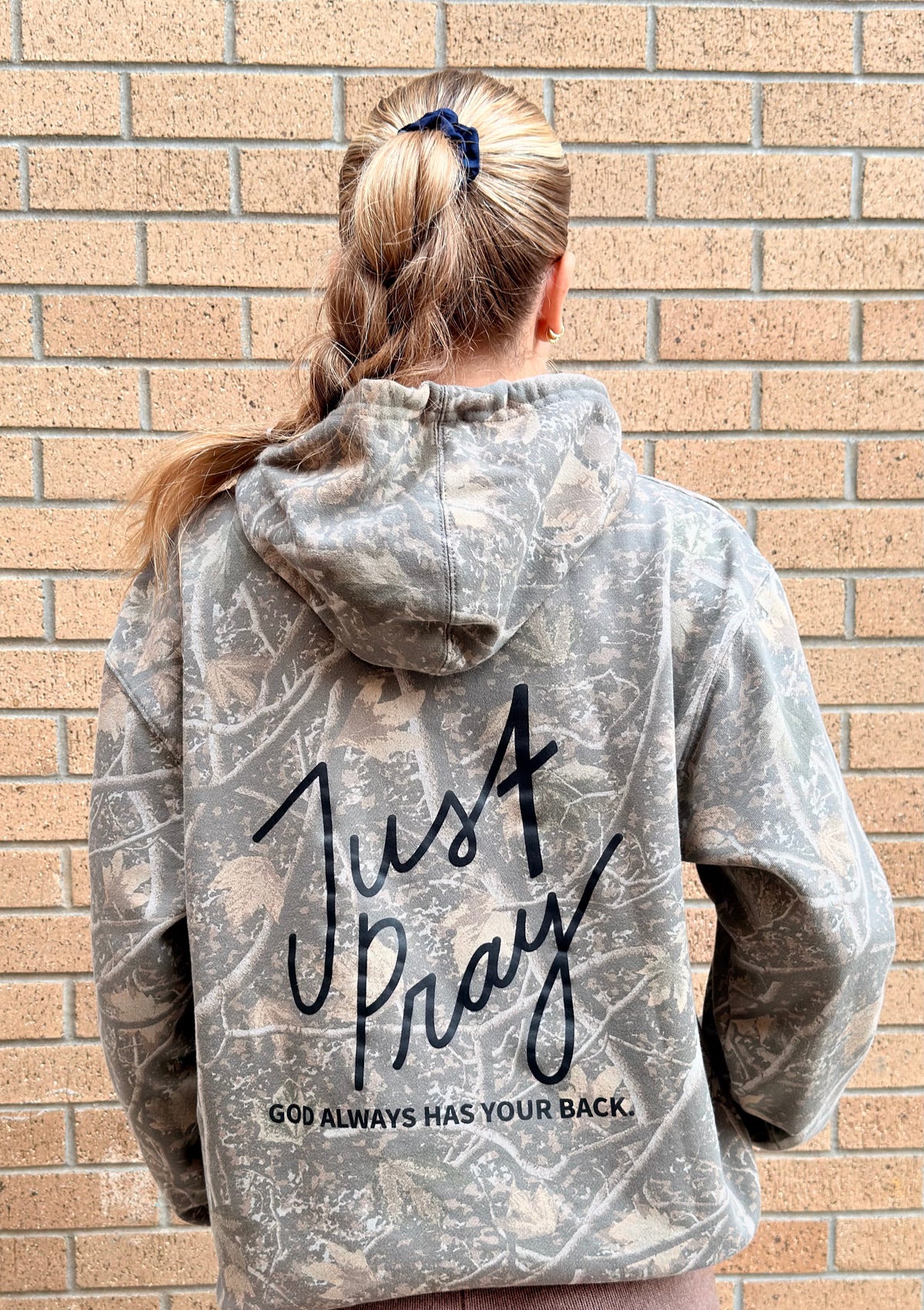 A woman viewed from behind wearing a gray and white distressed camouflage hoodie with 'Just Pray' and 'GOD ALWAYS HAS YOUR BACK.' printed in black on the back, standing against a tan brick wall.