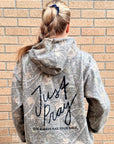 A woman viewed from behind wearing a gray and white distressed camouflage hoodie with 'Just Pray' and 'GOD ALWAYS HAS YOUR BACK.' printed in black on the back, standing against a tan brick wall.