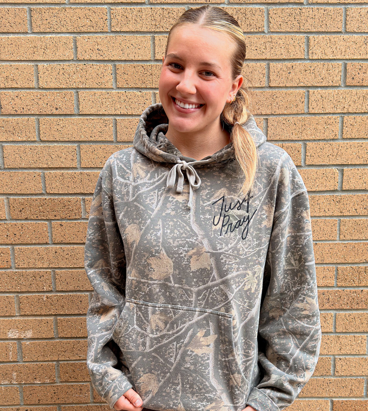 A smiling blonde woman wearing a gray camouflage hoodie with 'Just pray' printed in black script on the chest, standing against a tan brick wall.