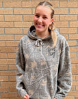A smiling blonde woman wearing a gray camouflage hoodie with 'Just pray' printed in black script on the chest, standing against a tan brick wall.