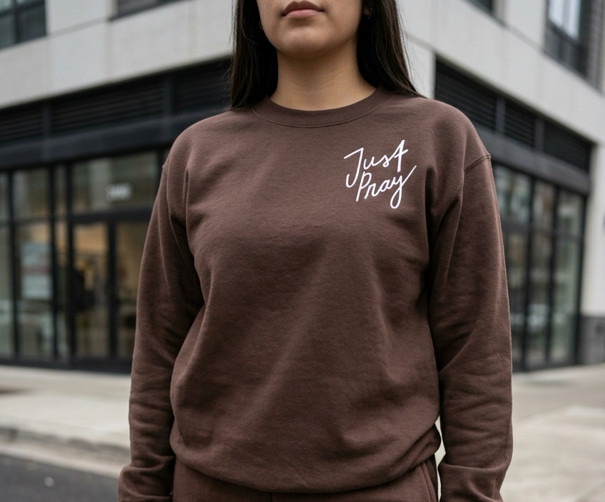 Person wearing a brown sweatshirt with 'Just Pray' text in white, standing in an urban setting.