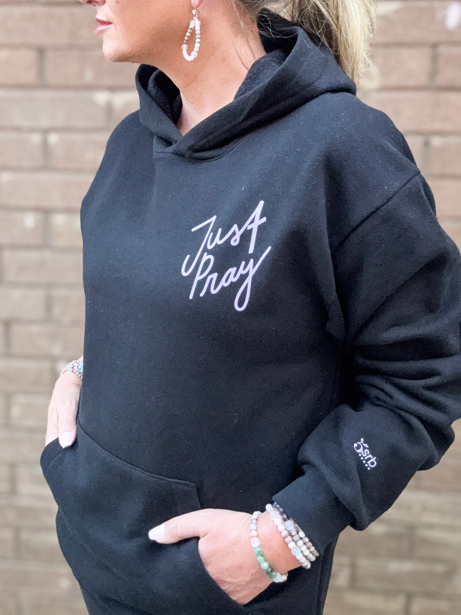 Person wearing a black hoodie with 'Just Pray' text, standing against a brick wall.
