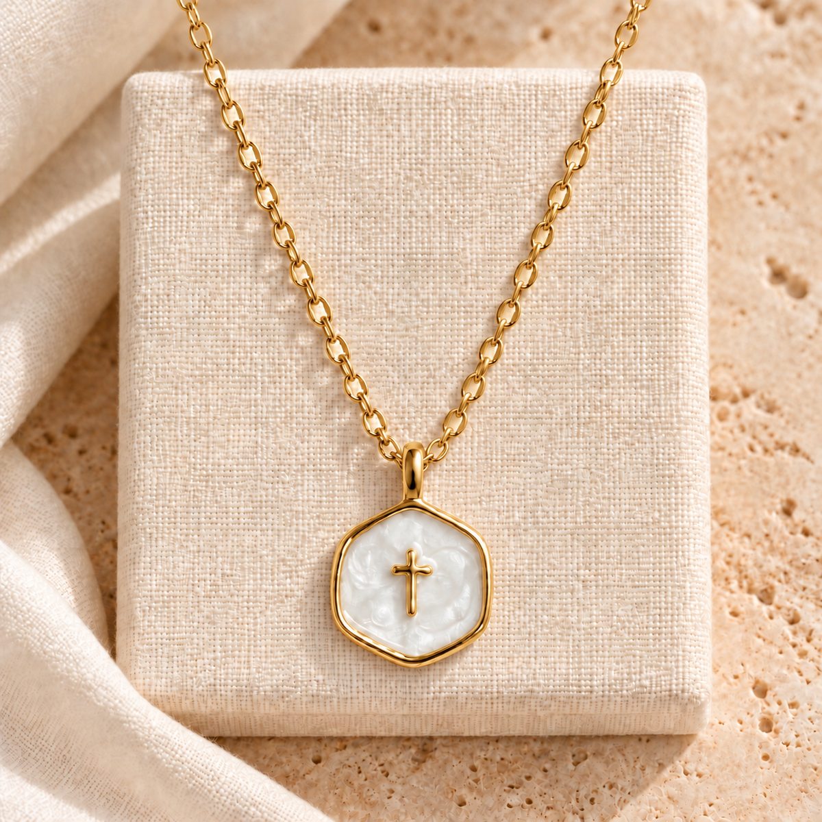 A gold chain necklace with a hexagonal pendant featuring a white mother-of-pearl cross, displayed on a textured beige jewelry box.