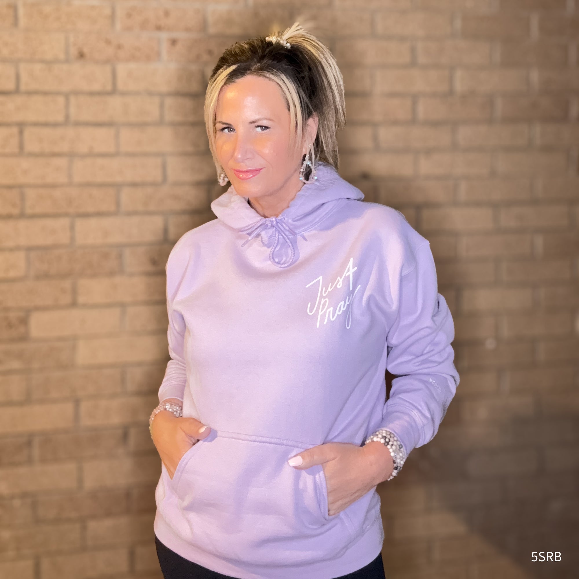 Woman wearing a light purple hoodie with text, standing against a brick wall.