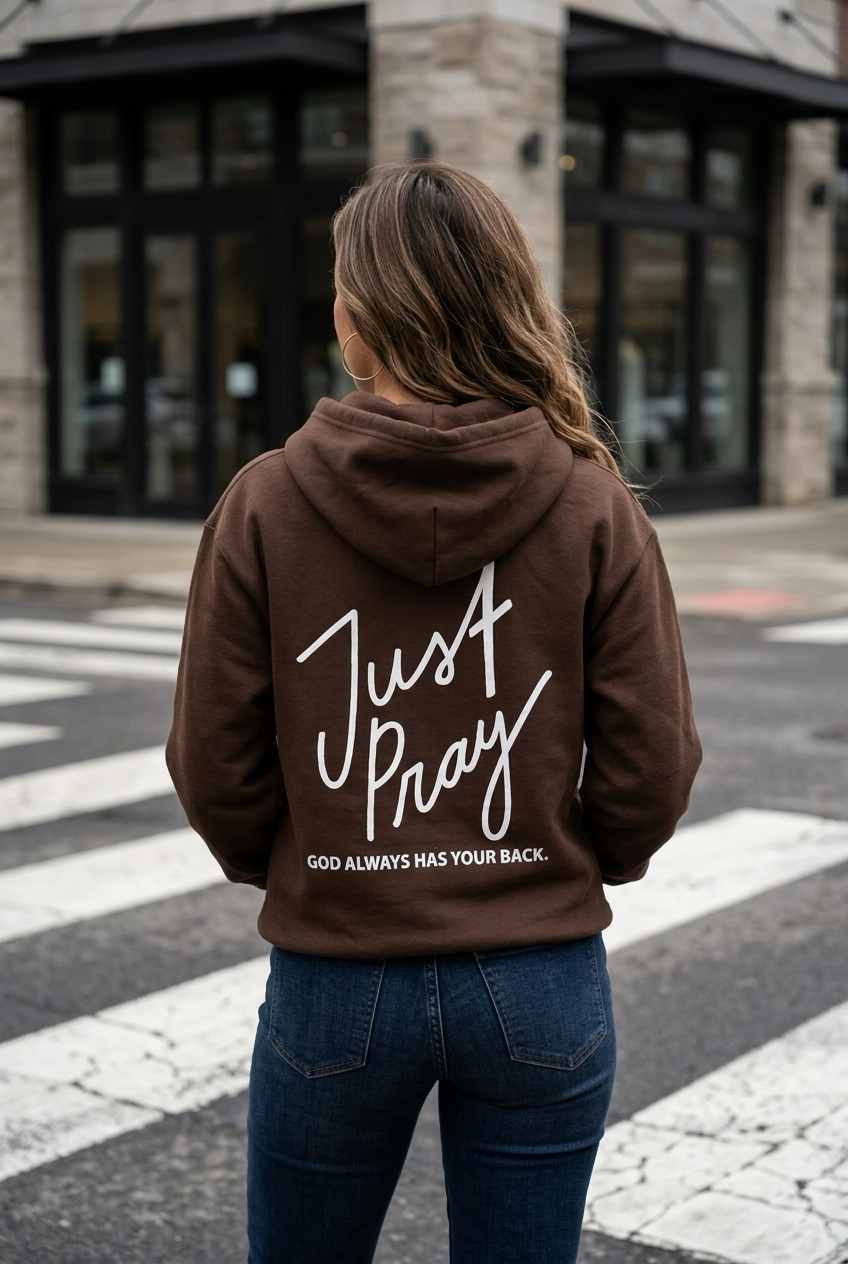 Person wearing a brown hoodie with 'Just Pray' text on the back, standing on a street.