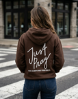 Person wearing a brown hoodie with 'Just Pray' text on the back, standing on a street.