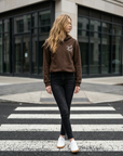 Woman walking on a crosswalk wearing a brown hoodie and black pants.
