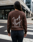 Person wearing a brown sweatshirt with 'Just Pray' text on the back, standing on a city street.
