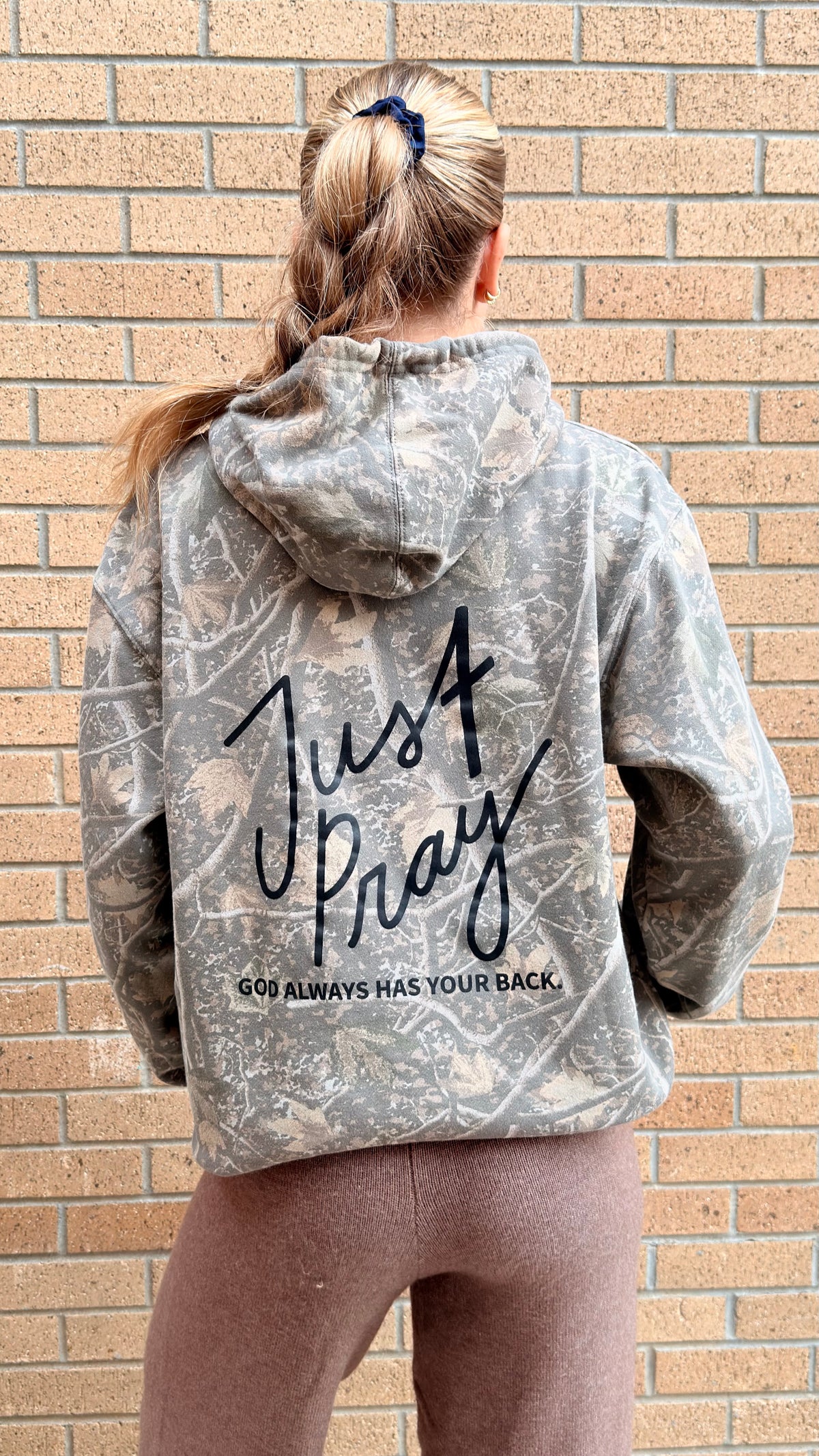 A woman viewed from behind wearing a gray and white distressed camouflage hoodie with 'Just Pray' and 'GOD ALWAYS HAS YOUR BACK.' printed in black on the back, standing against a tan brick wall.