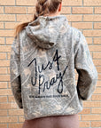 A woman viewed from behind wearing a gray and white distressed camouflage hoodie with 'Just Pray' and 'GOD ALWAYS HAS YOUR BACK.' printed in black on the back, standing against a tan brick wall.