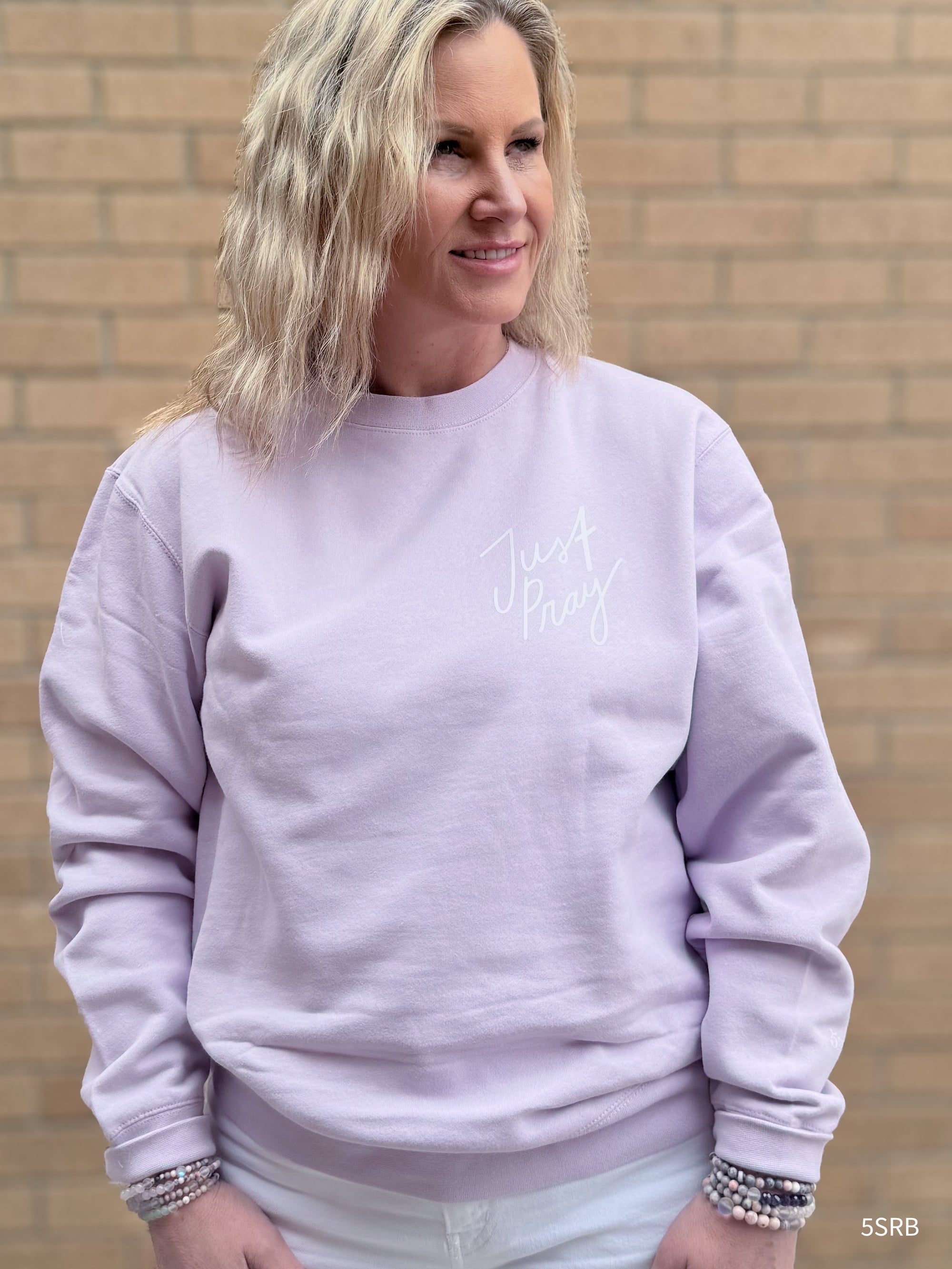 Woman wearing a light purple sweatshirt with text, standing against a brick wall.