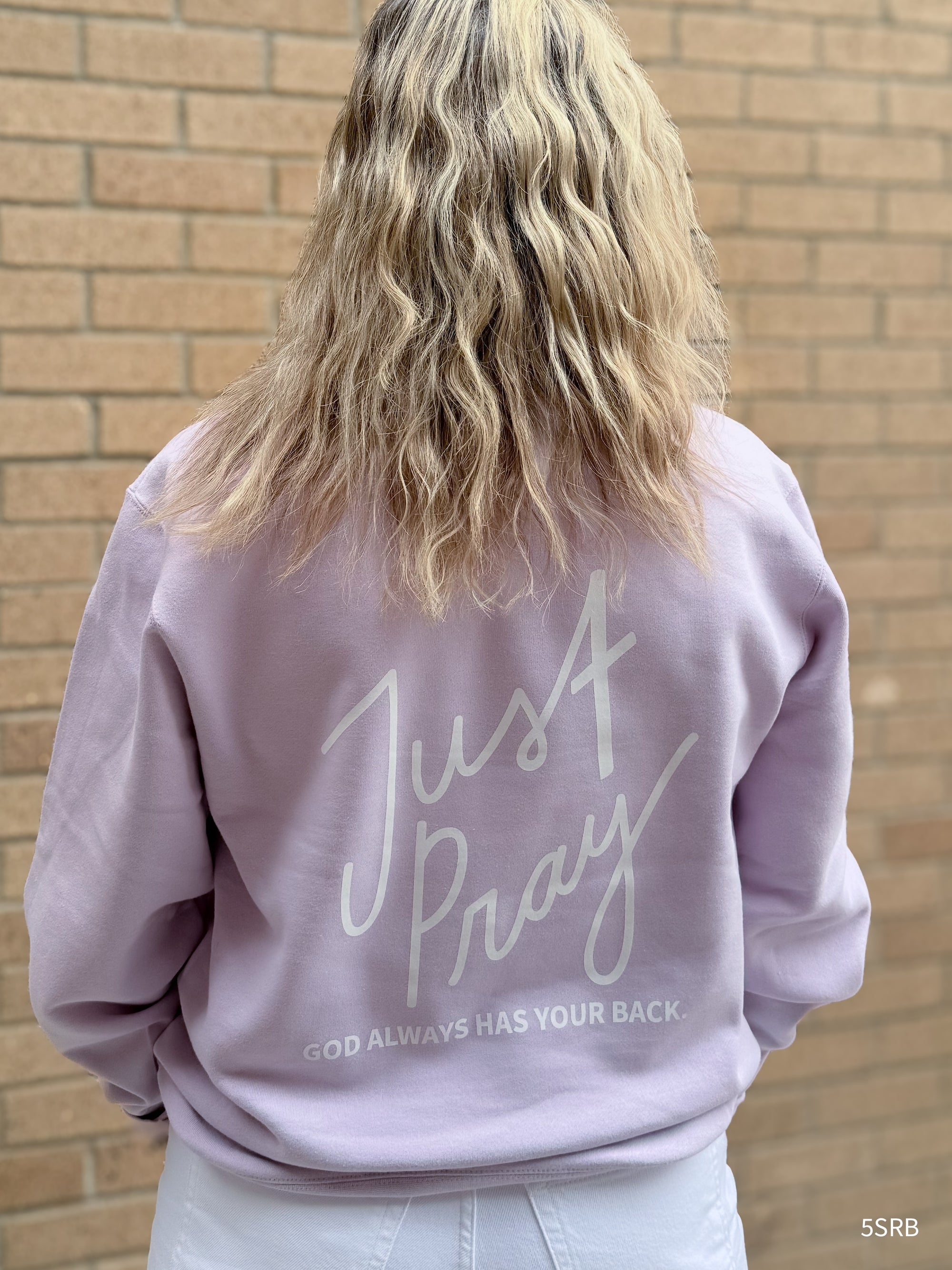 Person wearing a lavender sweatshirt with 'Just Pray' text against a brick wall.
