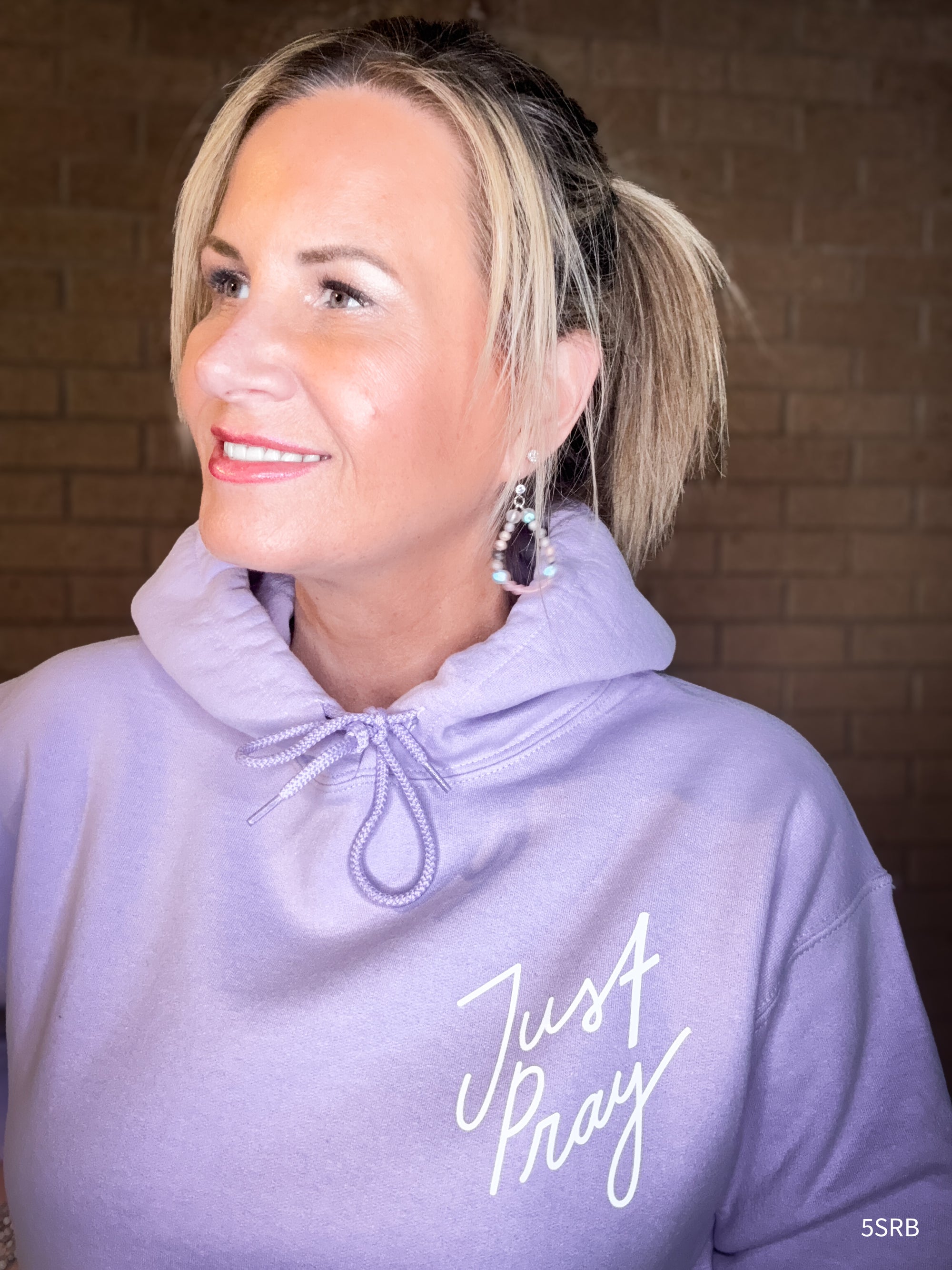 Woman wearing a lavender hoodie with 'Just Pray' text against a brick wall.
