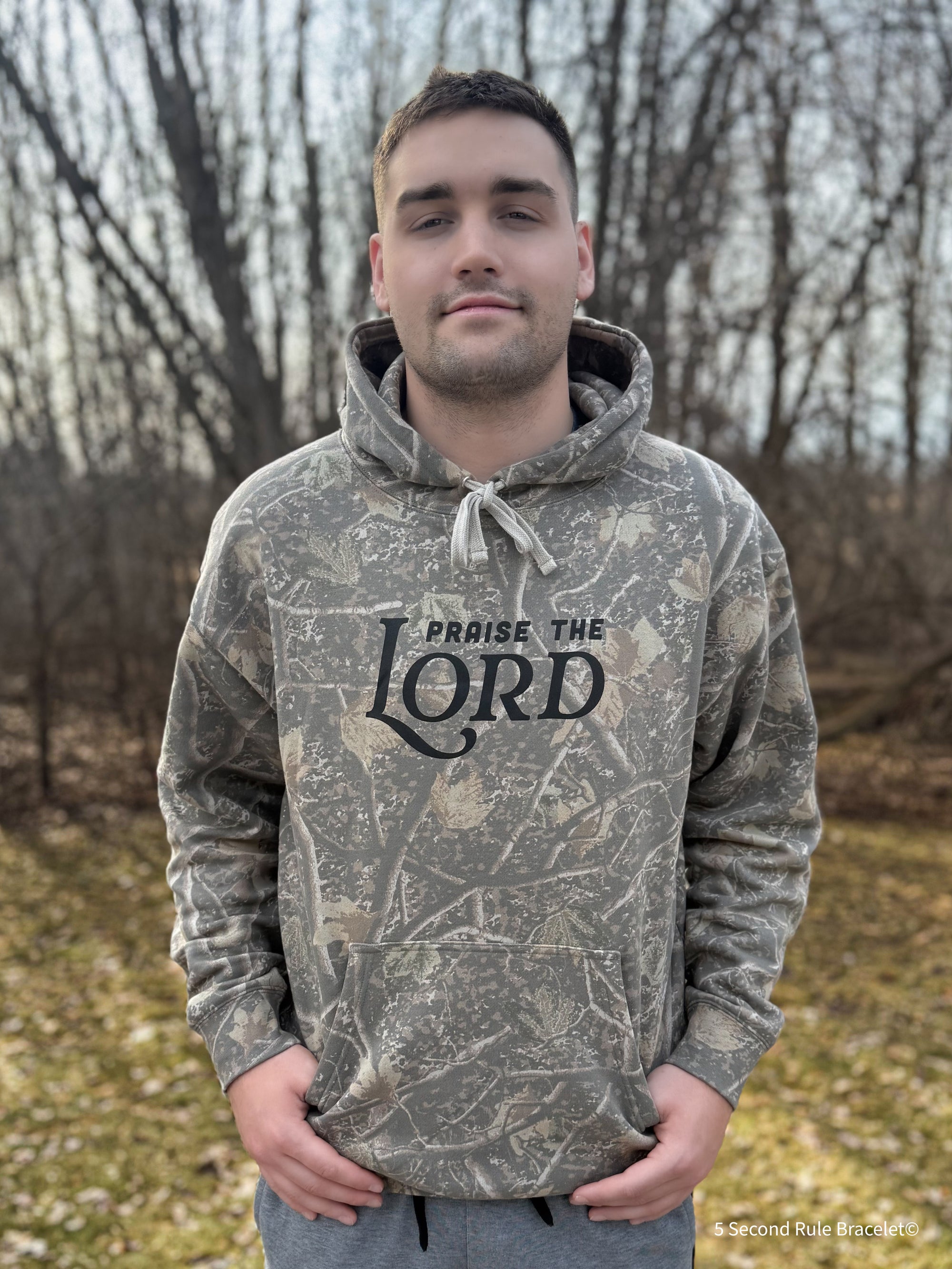 Person wearing a camouflage hoodie with 'Praise the Lord' text outdoors.