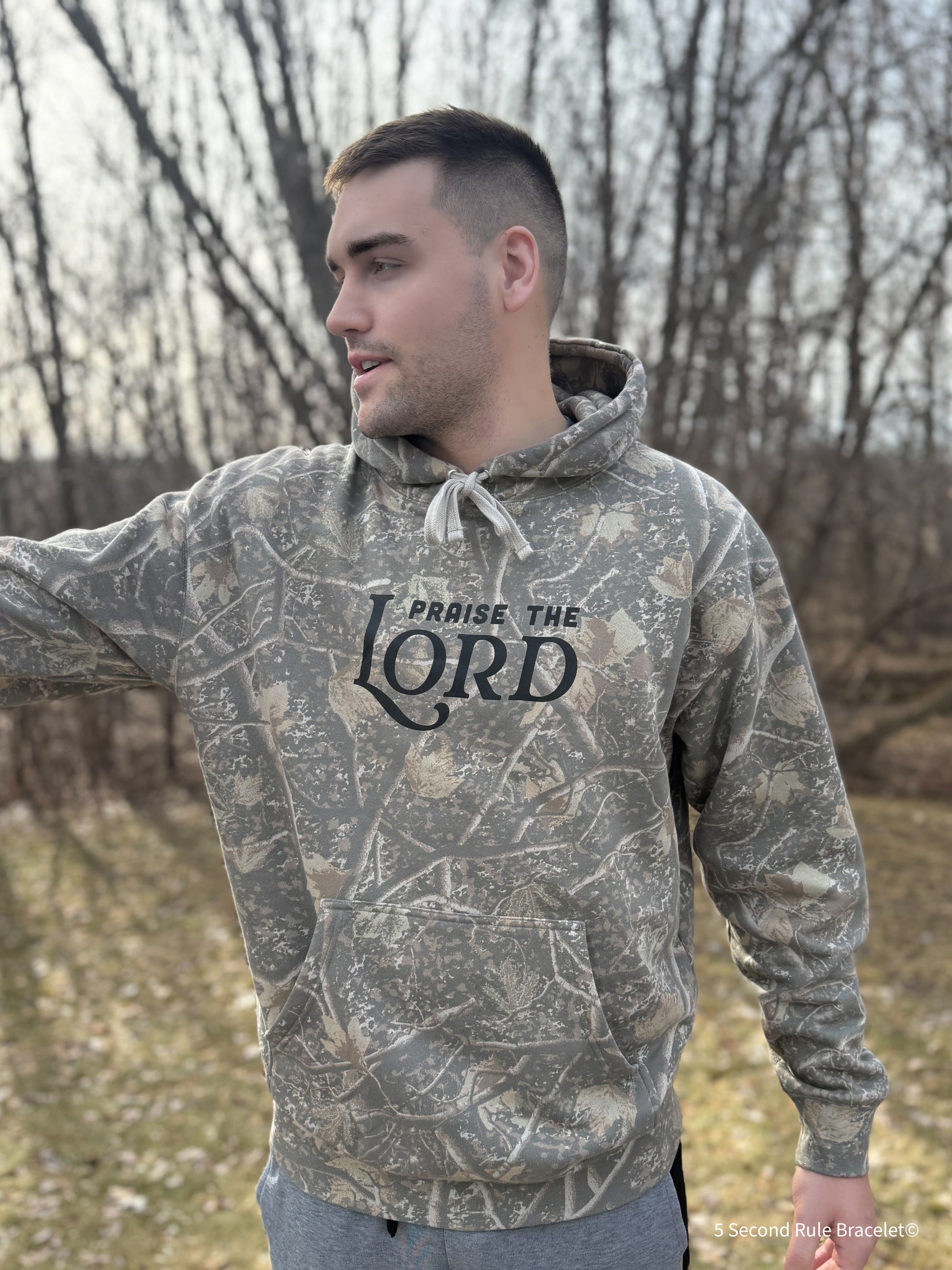 Man wearing a camouflage hoodie with 'Praise the Lord' text outdoors.