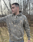 Man wearing a camouflage hoodie with 'Praise the Lord' text outdoors.