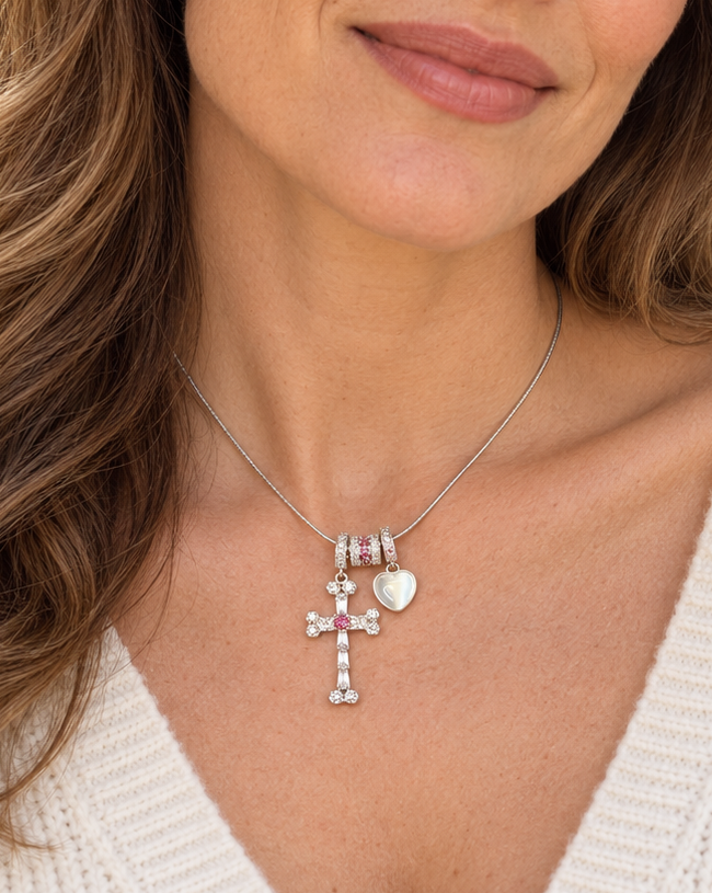 A close-up of a woman wearing a delicate silver cross necklace with pink gemstone accents and a small silver heart charm, paired with a white knit top.