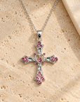 A silver cross pendant necklace adorned with pink gemstones, displayed on a textured beige stone surface.