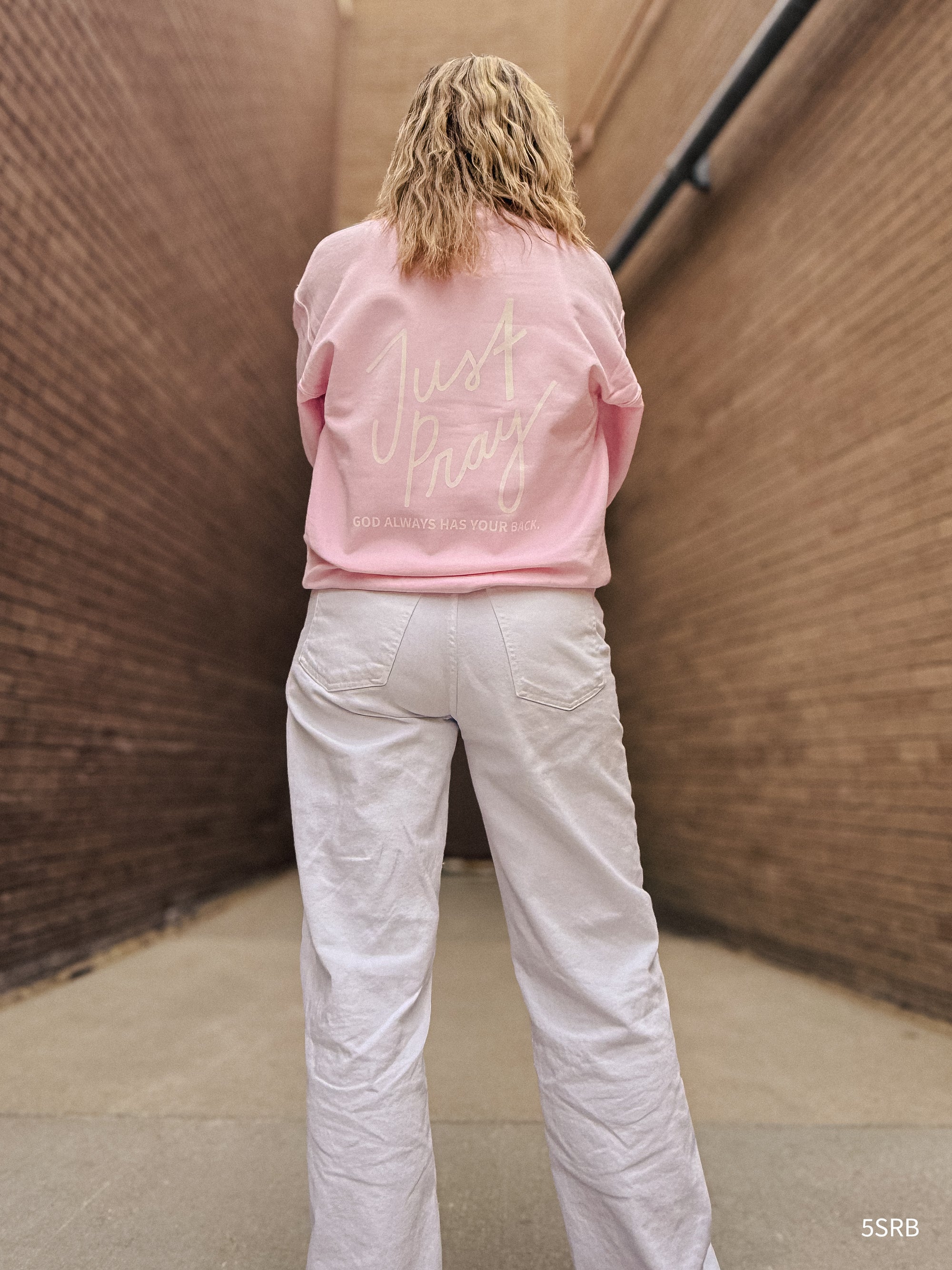 Person wearing a pink sweatshirt with text and light-colored pants against a textured wall.
