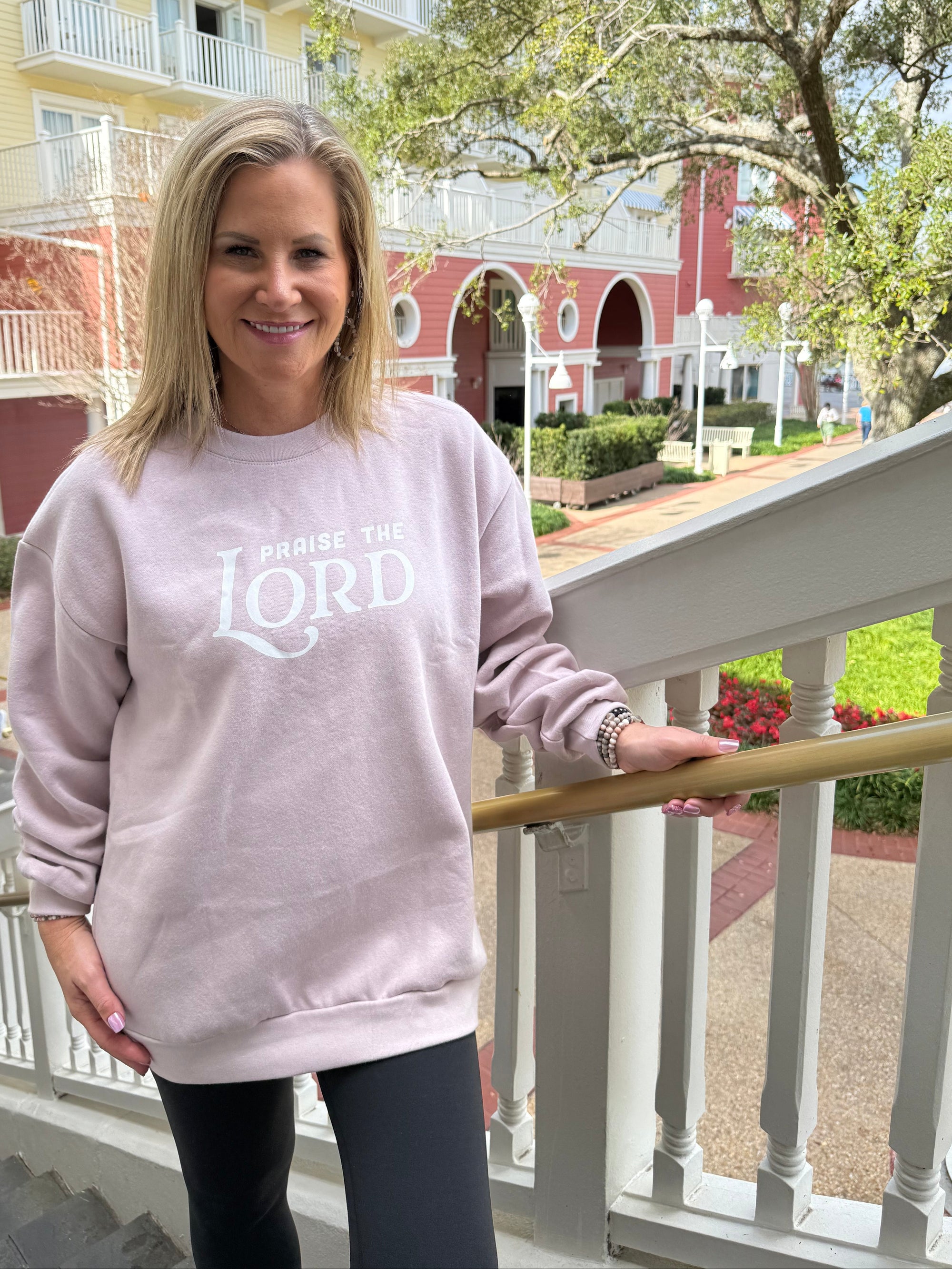 Woman wearing a pink sweatshirt with text, standing on a balcony with a building and trees in the background.