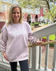 Woman wearing a pink sweatshirt with text, standing on a balcony with a building and trees in the background.